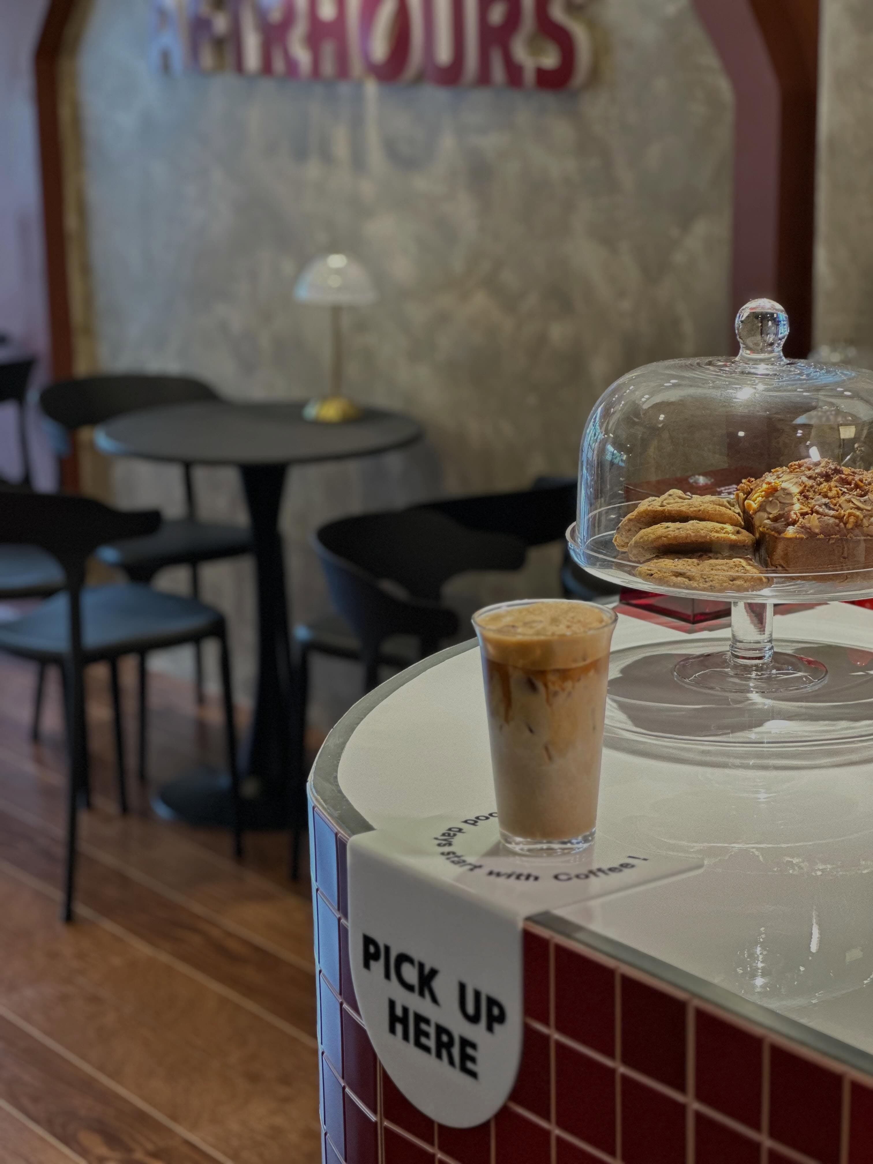 Iced coffee on the Aftrhours Cafe counter with pastries under a glass dome.